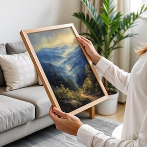 May include: A framed landscape painting of a mountain range with a winding road leading through a forest. The painting is in a wooden frame and is being held by a person.