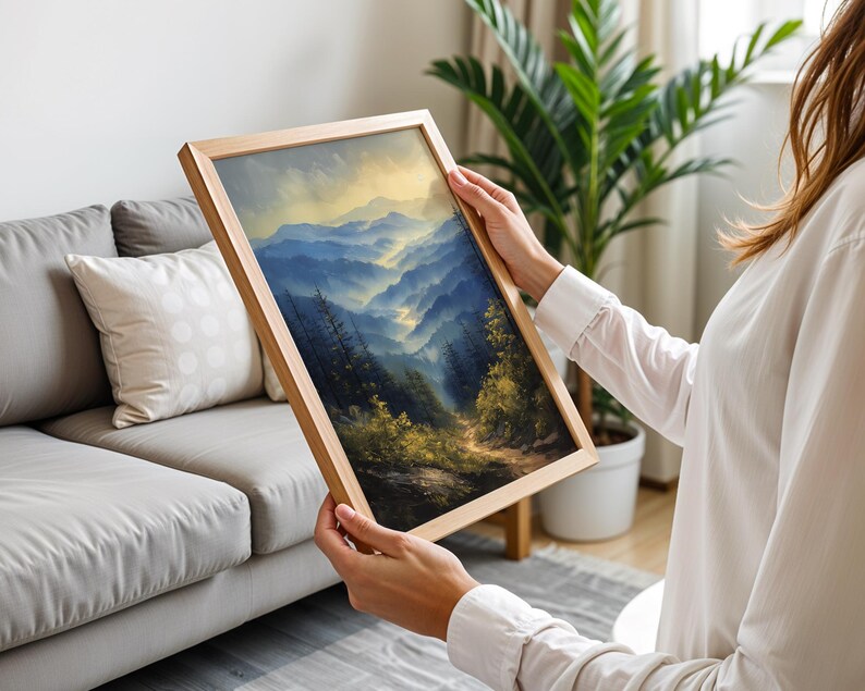 May include: A framed landscape painting of a mountain range with a winding road leading through a forest. The painting is in a wooden frame and is being held by a person.
