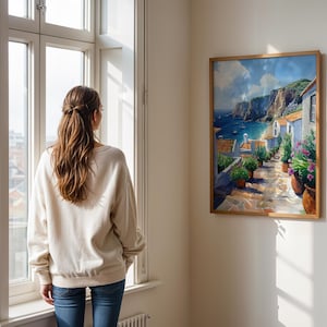 May include: A woman stands in front of a window looking at a painting of a coastal town with white buildings, a blue sea, and green hills. The painting is framed in wood and hangs on a white wall.