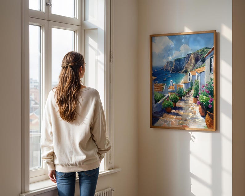 May include: A woman stands in front of a window looking at a painting of a coastal town with white buildings, a blue sea, and green hills. The painting is framed in wood and hangs on a white wall.