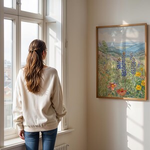 Puede incluir: Una pintura de acuarela enmarcada de un prado de flores silvestres con monta&ntilde;as azules en el fondo cuelga de una pared blanca. La pintura presenta flores coloridas en tonos de rojo, amarillo y azul. Una persona con una sudadera crema y vaqueros azules est&aacute; cerca de una ventana.