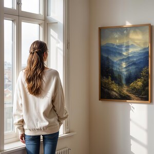 May include: A woman stands in front of a window looking at a framed landscape painting of a mountain range with a path leading through the trees. The painting is in a wooden frame and hangs on a white wall.
