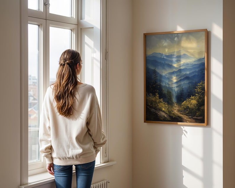 May include: A woman stands in front of a window looking at a framed landscape painting of a mountain range with a path leading through the trees. The painting is in a wooden frame and hangs on a white wall.