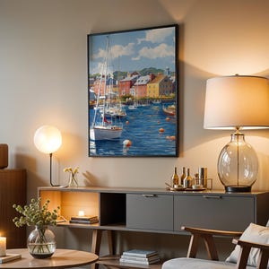 May include: A framed art print depicts a harbor scene with sailboats and colorful buildings. The artwork features a blue sea, white sailboats, and buildings with red, yellow, and brown facades. The print is displayed in a living room setting.