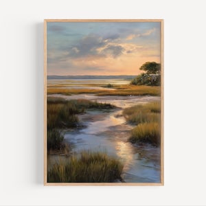 May include: A framed landscape painting shows a marsh scene at sunset. The artwork features a waterway with tall grasses, a tree, and a colorful sky with shades of orange, blue, and yellow. The frame is made of light-coloured wood.
