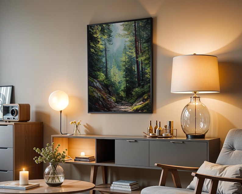 May include: A living room scene featuring a framed forest landscape painting, a table lamp with a glass base, and a modern gray cabinet. A round table holds a vase with greenery and a lit candle. The room is bathed in warm light.