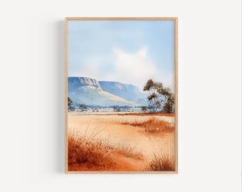 May include: A framed watercolor painting depicting a landscape with a vast golden-brown field in the foreground, sparse trees, and a distant blue mountain range under a pale sky with soft white clouds, all enclosed in a light wood frame.