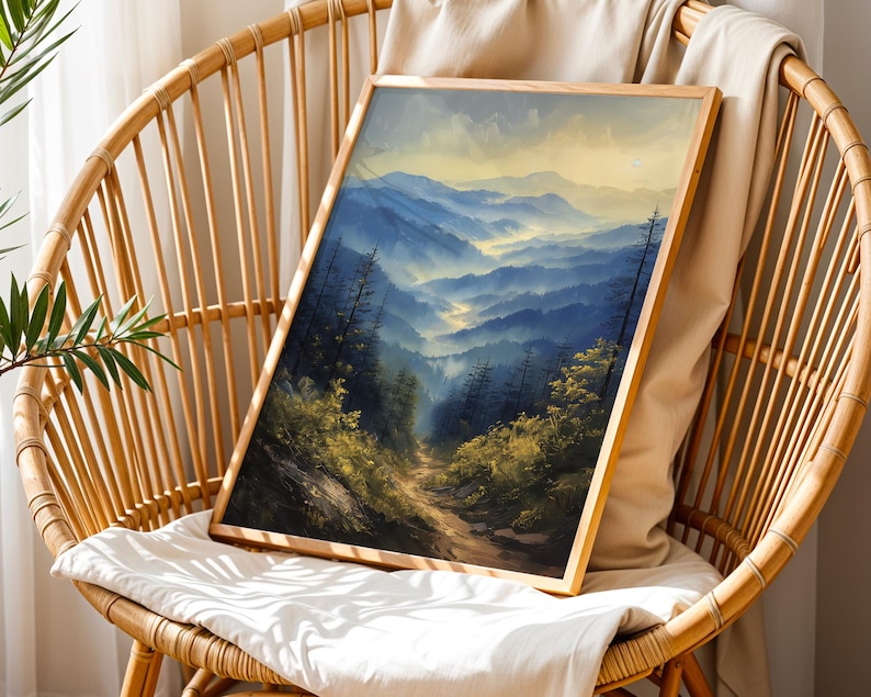 May include: A framed painting of a mountain landscape with a winding path leading through the trees. The mountains are painted in shades of blue and green, with a hazy mist in the distance. The painting is set against a backdrop of a light brown wicker chair.