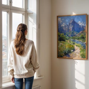 May include: A woman stands in front of a window looking at a framed painting of a mountain landscape with a river and a path leading through the scene.