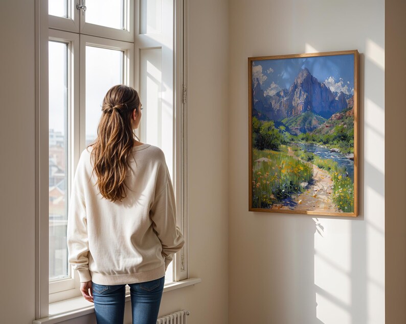 May include: A woman stands in front of a window looking at a framed painting of a mountain landscape with a river and a path leading through the scene.