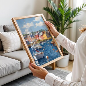 May include: A framed art print depicting a harbor scene with sailboats, buildings, and blue water. The artwork is in a wooden frame and features a bright, colorful palette. The print is being held up by a person.