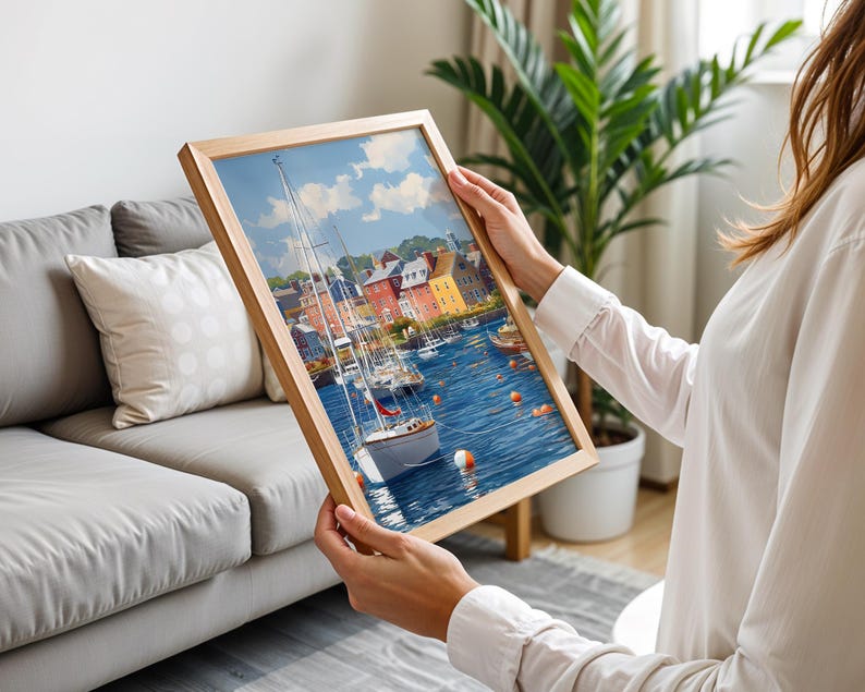 May include: A framed art print depicting a harbor scene with sailboats, buildings, and blue water. The artwork is in a wooden frame and features a bright, colorful palette. The print is being held up by a person.
