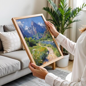 May include: A framed landscape painting of a mountain range with a river flowing through a valley. The painting is in a wooden frame and is being held by a person.