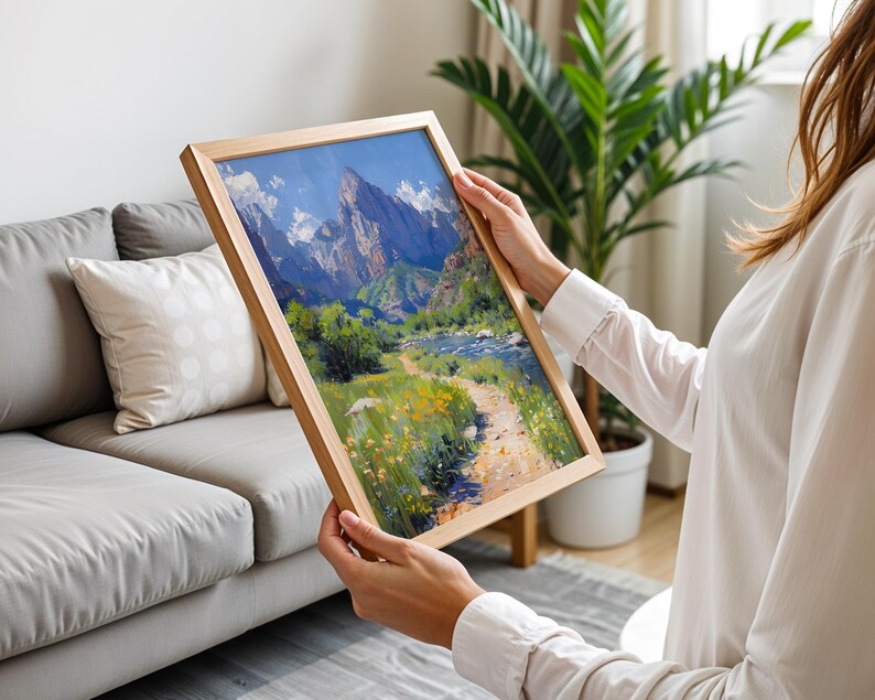 May include: A framed landscape painting of a mountain range with a river flowing through a valley. The painting is in a wooden frame and is being held by a person.
