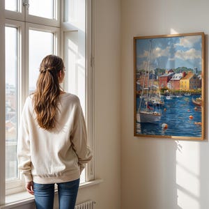 May include: A framed nautical painting depicts a harbor scene with sailboats, buildings, and a blue sea. The artwork is displayed on a wall next to a window. A person wearing a cream-colored sweatshirt and blue jeans is looking out the window.