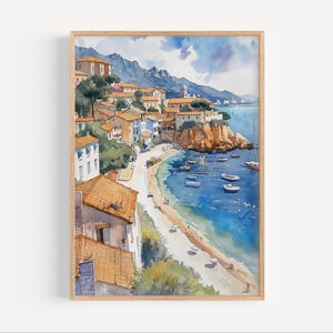 May include: Watercolor painting of a coastal town with buildings featuring orange tile roofs, set against a backdrop of mountains and a blue sea. Small boats dot the water, and a sandy beach lines the shore. The artwork is framed in a light-colored wooden frame.
