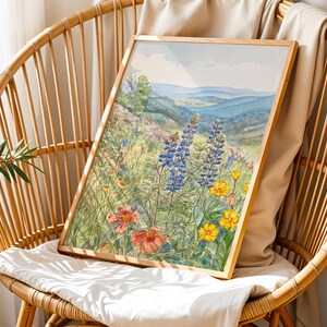 Puede incluir: Una impresi&oacute;n en acuarela enmarcada de un prado de flores silvestres con flores azules, amarillas y rojas. La obra de arte representa un paisaje con colinas y un cielo azul. La impresi&oacute;n se muestra en una silla de mimbre.