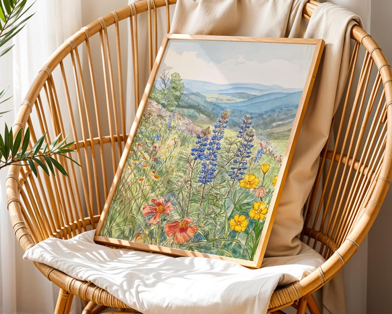 Puede incluir: Una impresi&oacute;n en acuarela enmarcada de un prado de flores silvestres con flores azules, amarillas y rojas. La obra de arte representa un paisaje con colinas y un cielo azul. La impresi&oacute;n se muestra en una silla de mimbre.