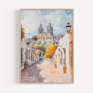 May include: Watercolour painting of a European street scene. The artwork depicts a sunlit cobbled street lined with white buildings and orange accents. A large cathedral is visible in the background under a blue sky. The painting is framed in light wood.