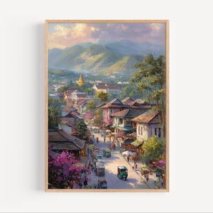 May include: A framed landscape painting depicts a vibrant street scene in a Southeast Asian town. The artwork features buildings with brown roofs, lush greenery, and a golden pagoda. The sky transitions from pink to blue.
