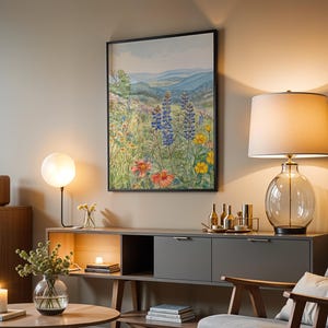 Puede incluir: Una escena de sal&oacute;n con una pintura de acuarela enmarcada de un prado de flores silvestres. Una l&aacute;mpara de cristal con una pantalla blanca y una base de cristal transparente se encuentra sobre un mueble gris. Una mesa redonda tiene una vela y un jarr&oacute;n de vegetaci&oacute;n.