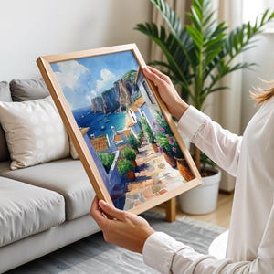 May include: A framed painting of a coastal town with colorful buildings, a winding cobblestone street, and a view of the ocean. The painting is in a wooden frame and is being held by a person.