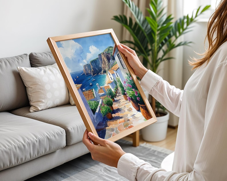 May include: A framed painting of a coastal town with colorful buildings, a winding cobblestone street, and a view of the ocean. The painting is in a wooden frame and is being held by a person.