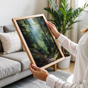 May include: A framed landscape painting depicting a lush forest scene with tall trees and a winding path. The artwork features a palette of greens, browns, and blues, creating a sense of depth and tranquility. The frame is made of light-colored wood.