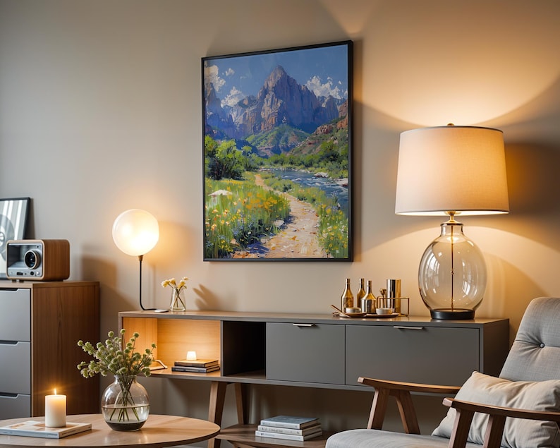 May include: A living room interior with a gray couch, a wooden coffee table, a side table with a lamp, and a large framed painting of a mountain landscape with a river running through it.