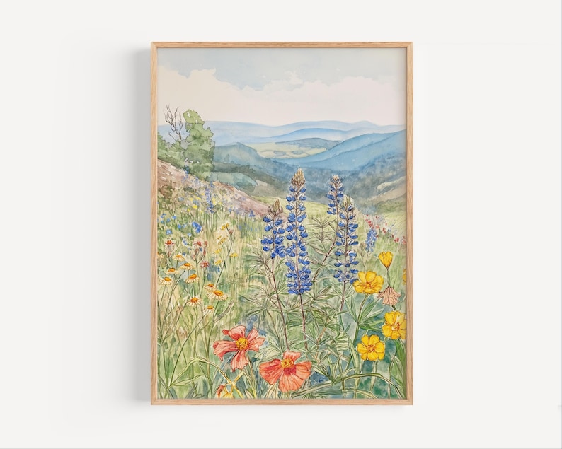 Puede incluir: Una impresi&oacute;n en acuarela enmarcada de un prado de flores silvestres con un tel&oacute;n de fondo de monta&ntilde;a. La obra de arte presenta altramuces azules vibrantes, flores silvestres naranjas y amarillas y colinas onduladas en tonos azules y verdes. La impresi&oacute;n est&aacute; en un marco de madera natural.