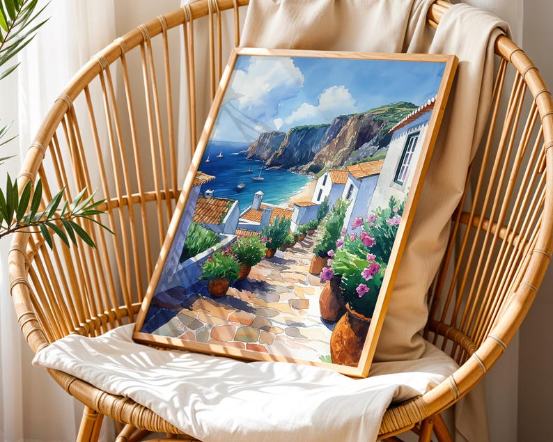May include: A watercolor painting of a coastal village with white houses, a blue sea, and green hills. The painting is framed in a wooden frame and is sitting on a wicker chair.