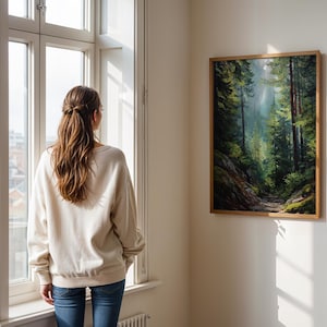 May include: A framed landscape painting of a forest scene with tall trees and a path. The painting is displayed on a white wall. A person wearing a cream-colored sweatshirt and blue jeans stands near a window, looking at the painting.