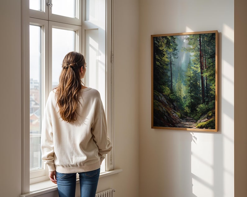 May include: A framed landscape painting of a forest scene with tall trees and a path. The painting is displayed on a white wall. A person wearing a cream-colored sweatshirt and blue jeans stands near a window, looking at the painting.