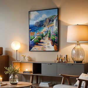 May include: A framed painting of a coastal village with white houses, a blue ocean, and a rocky cliff. The painting is hanging on a wall in a living room with a lamp, a side table, and a chair.