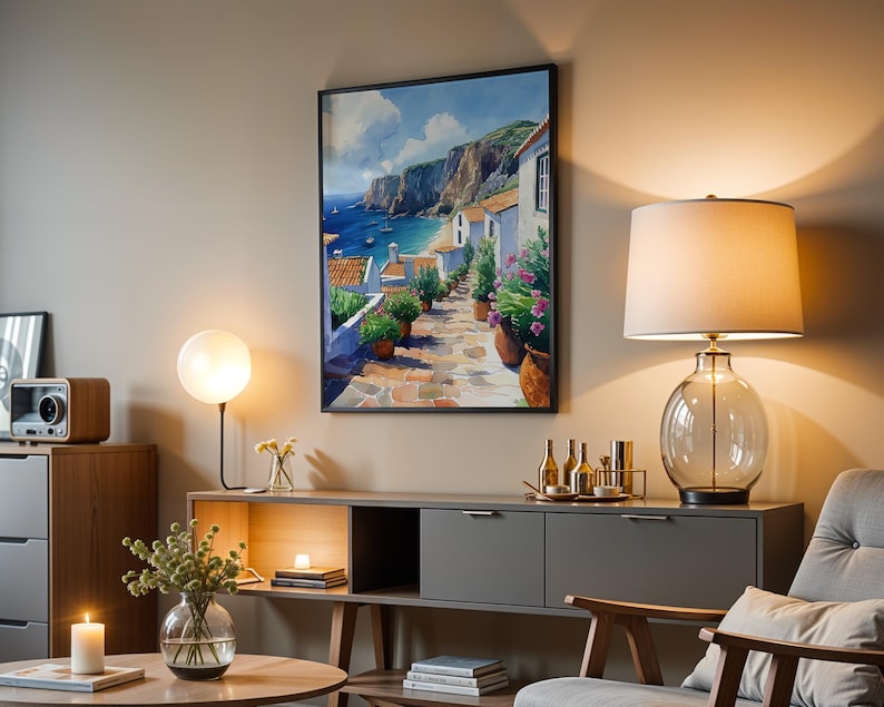 May include: A framed painting of a coastal village with white houses, a blue ocean, and a rocky cliff. The painting is hanging on a wall in a living room with a lamp, a side table, and a chair.