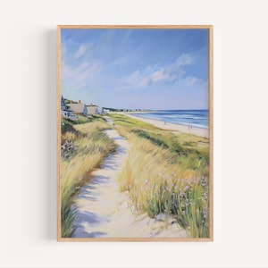 Puede incluir: Pintura de paisaje enmarcada de una escena de playa. Un sendero arenoso conduce a través de altas hierbas doradas hacia el océano bajo un cielo azul con nubes. Casas blancas bordean la costa. La obra de arte está en un marco de madera clara.
