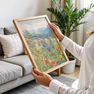 Puede incluir: Una pintura de acuarela enmarcada de un prado de flores silvestres con monta&ntilde;as azules al fondo. La obra presenta coloridas flores silvestres en tonos de rojo, amarillo y azul, enmarcada en madera natural.