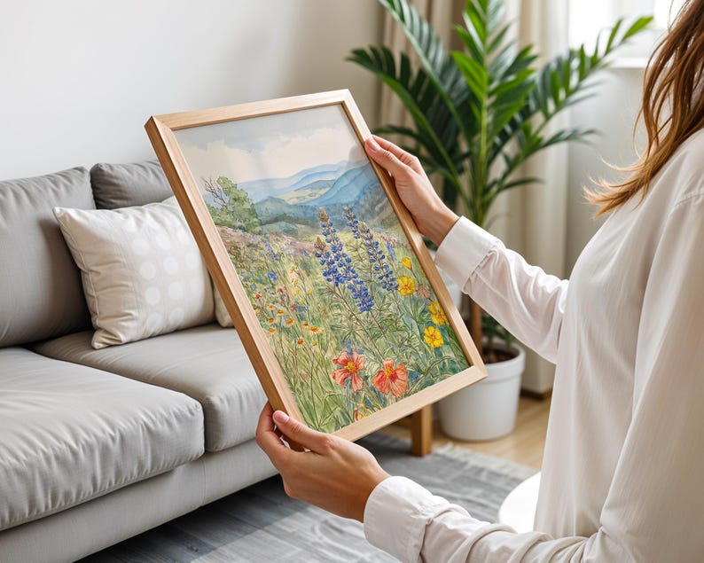 Puede incluir: Una pintura de acuarela enmarcada de un prado de flores silvestres con monta&ntilde;as azules al fondo. La obra presenta coloridas flores silvestres en tonos de rojo, amarillo y azul, enmarcada en madera natural.