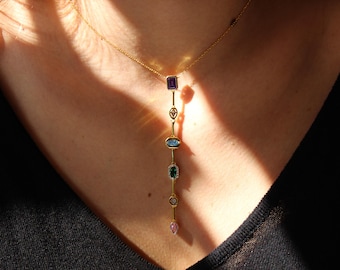 May include: A gold chain necklace with a long pendant featuring five colorful gemstones. The pendant has a teardrop-shaped gemstone at the bottom, followed by a round gemstone, an oval gemstone, a rectangular gemstone, and a square gemstone at the top.