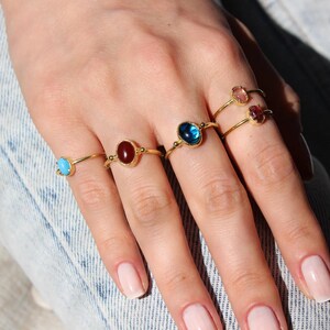 Op de afbeelding: Een hand draagt vijf gouden ringen met verschillende edelstenen. De ringen hebben ovale stenen in de kleuren blauw, rood, roze en turquoise.