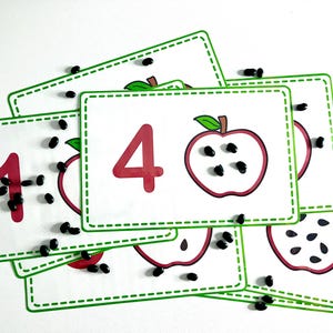 Op de afbeelding: Een set van vijf witte kaarten met groene randen en zwarte cijfers. Elke kaart heeft een rode appel met zwarte pitten. De kaarten liggen in een stapel, met de kaart met het cijfer 4 bovenop. Er liggen zwarte pitten verspreid rond de kaarten.