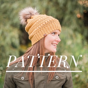 May include: A crocheted beanie hat with a faux fur pom pom in a mustard yellow color. The hat has a textured stitch pattern and is worn by a person with long brown hair. The word "PATTERN" is written in white text across the bottom of the image.