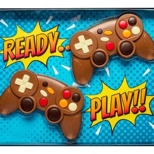 Puede incluir: Dos mandos de videojuegos de chocolate marrón con detalles blancos, rojos y naranjas se muestran en una caja azul. Las palabras "READY" y "PLAY!!" están en bocadillos amarillos.