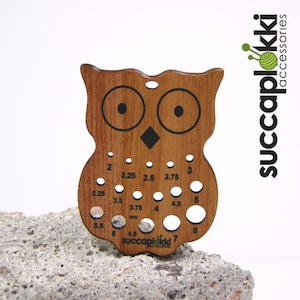 May include: Wooden owl-shaped knitting needle gauge with a black and white owl design and holes for different needle sizes, labelled in millimetres and US sizes. The gauge is made by Succaplokki Accessories.