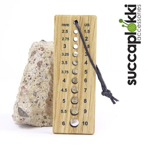 May include: Wooden knitting needle gauge with a black cord. The gauge has markings for both US and metric sizes. The gauge is labeled "succaplokki accessories".