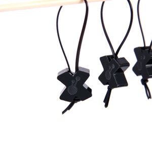 Könnte beinhalten: Nahaufnahme mehrerer schwarzer Maschenmarkierer, die an einem Holzstab hängen. Jeder Marker ist X-förmig mit einem kleinen, detaillierten Design und einer Schlaufe aus schwarzem Kordel. Die Marker sind vor weißem Hintergrund.