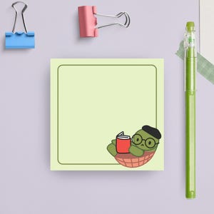 May include: A green turtle wearing a beret and glasses is reading a red book while relaxing in a shell. The turtle is on a light green sticky note with a dark green border.