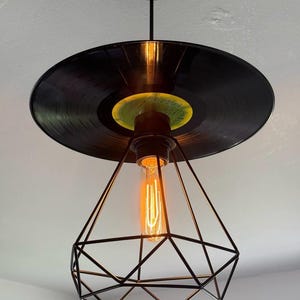 May include: A black vinyl record is used as the ceiling fixture for a pendant light. The light bulb is encased in a black geometric cage.