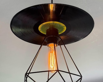 May include: A black vinyl record is used as the ceiling fixture for a pendant light. The light bulb is encased in a black geometric cage.