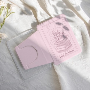 May include: A light pink phone stand with a clear phone case. The stand features a tarot card design with a floral illustration and the text "THE READER". The words "happylifeenjoydesign" are at the bottom.